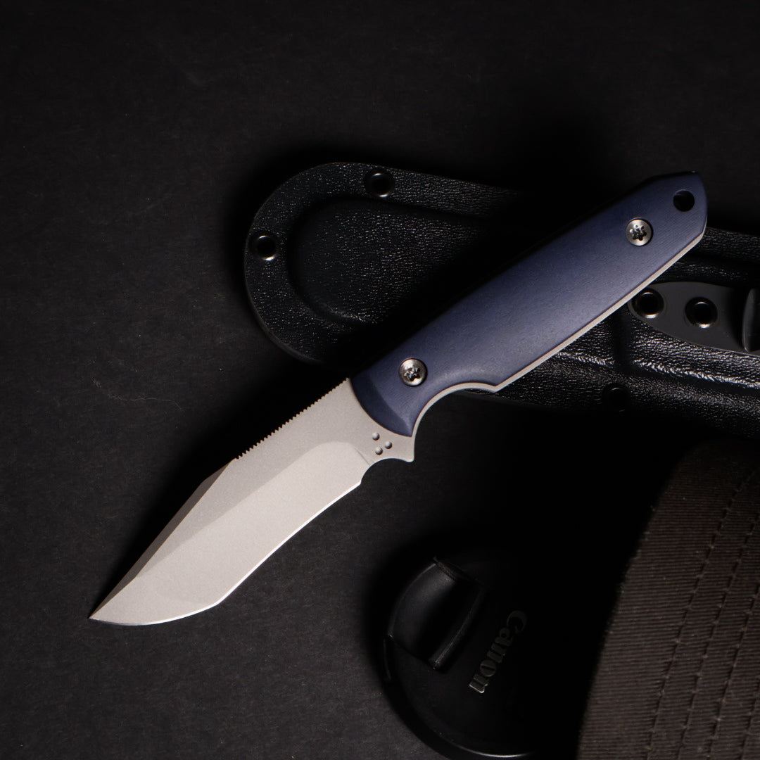 fixed blade knife with blue handle resting on kydex sheath next to camera lens cover and ballcap on black surface.