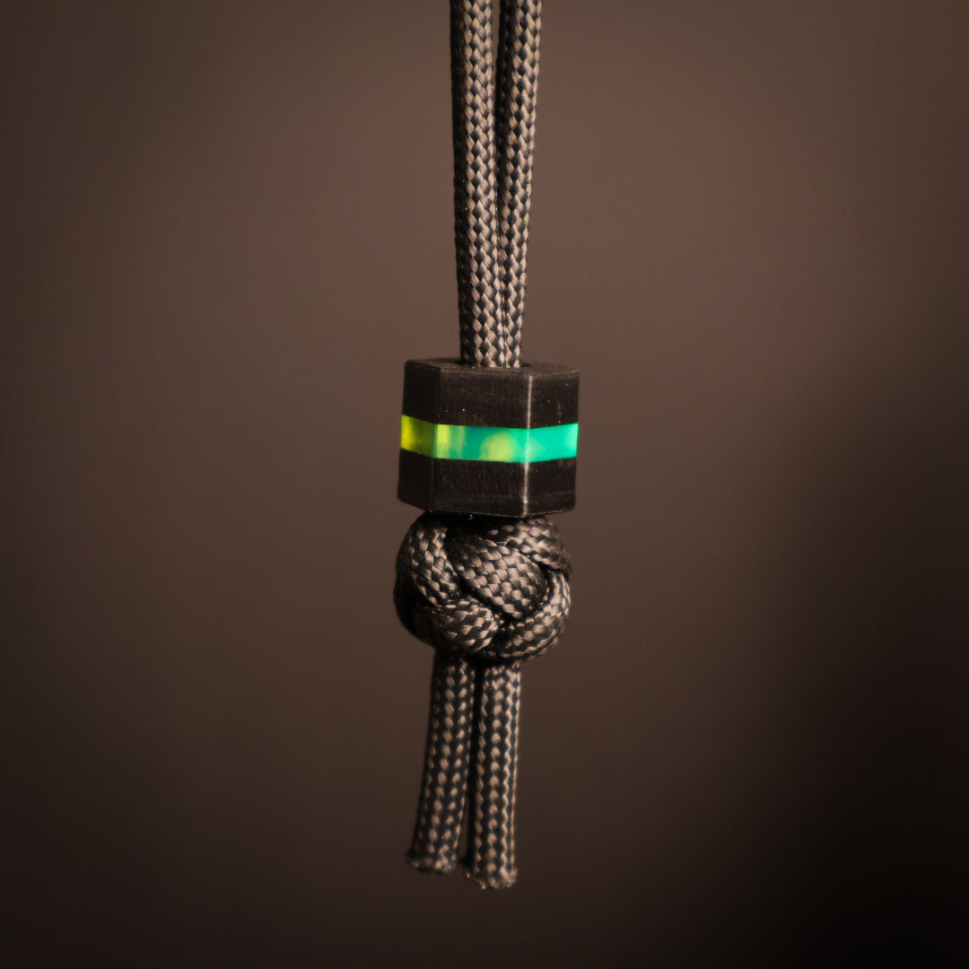 Hexagonal lanyard bead with fluorescent green layer sandwiched between black layers. Hanging on paracord lanyard