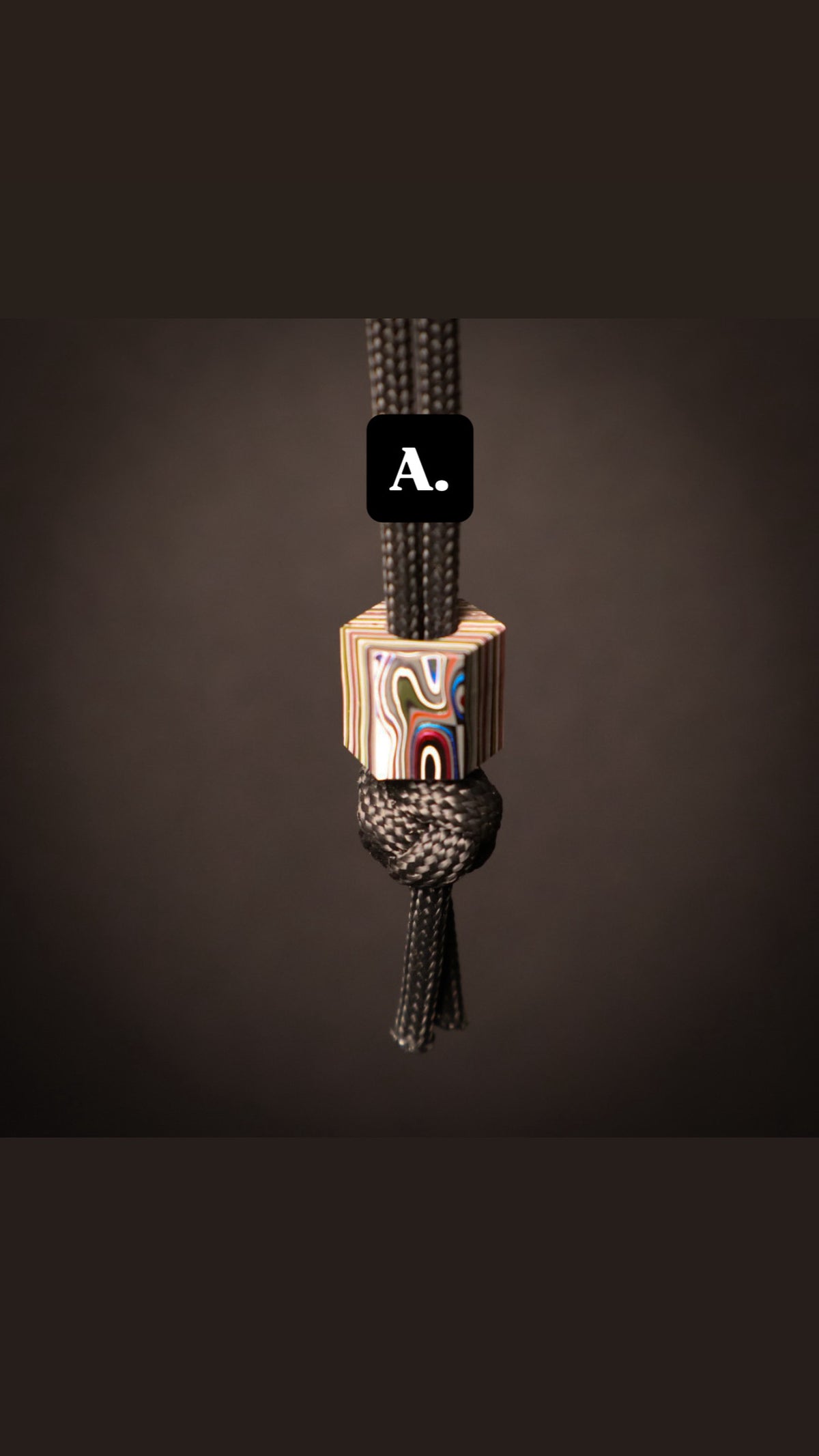 A close-up of a black braided cord with a knotted section and a small decorative wooden bead featuring an abstract multicolor pattern. A black label with the white text 'A.