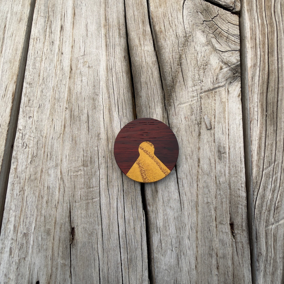 A round wooden edc coin with a dark reddish-brown top half and a gold-toned lower half placed on a weathered gray wooden plank surface.