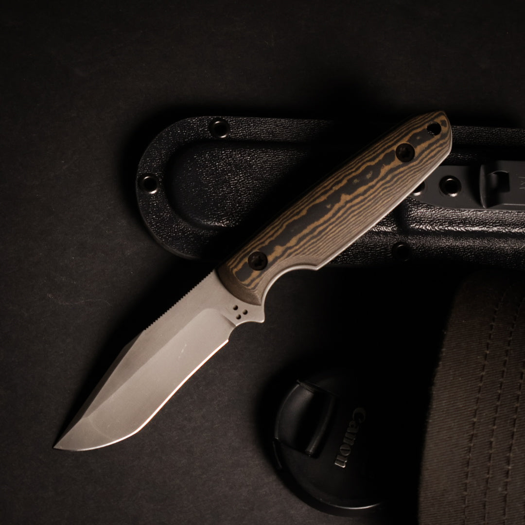 fixed blade knife with brown and black handle resting on kydex sheath next to camera lens cover and ballcap on black surface. 
