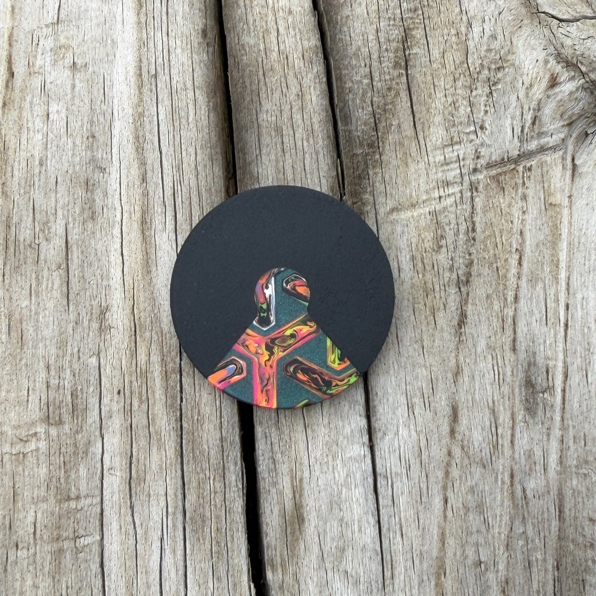 A round black coin with a triangular cutout with a colorful marbled piece inlaid, placed on a rustic weathered wood background.
