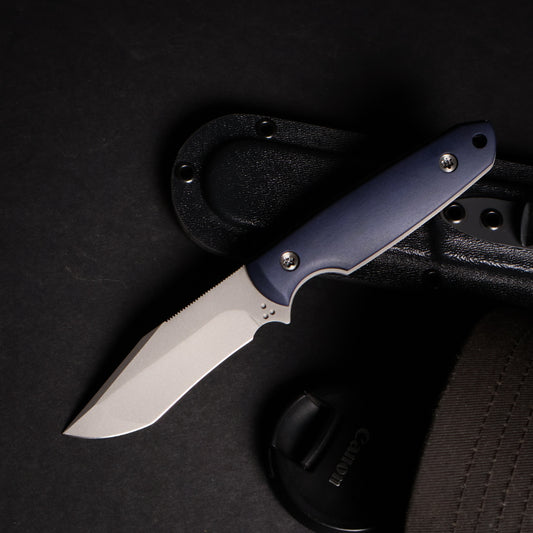 fixed blade knife with blue handle resting on kydex sheath next to camera lens cover and ballcap on black surface. 