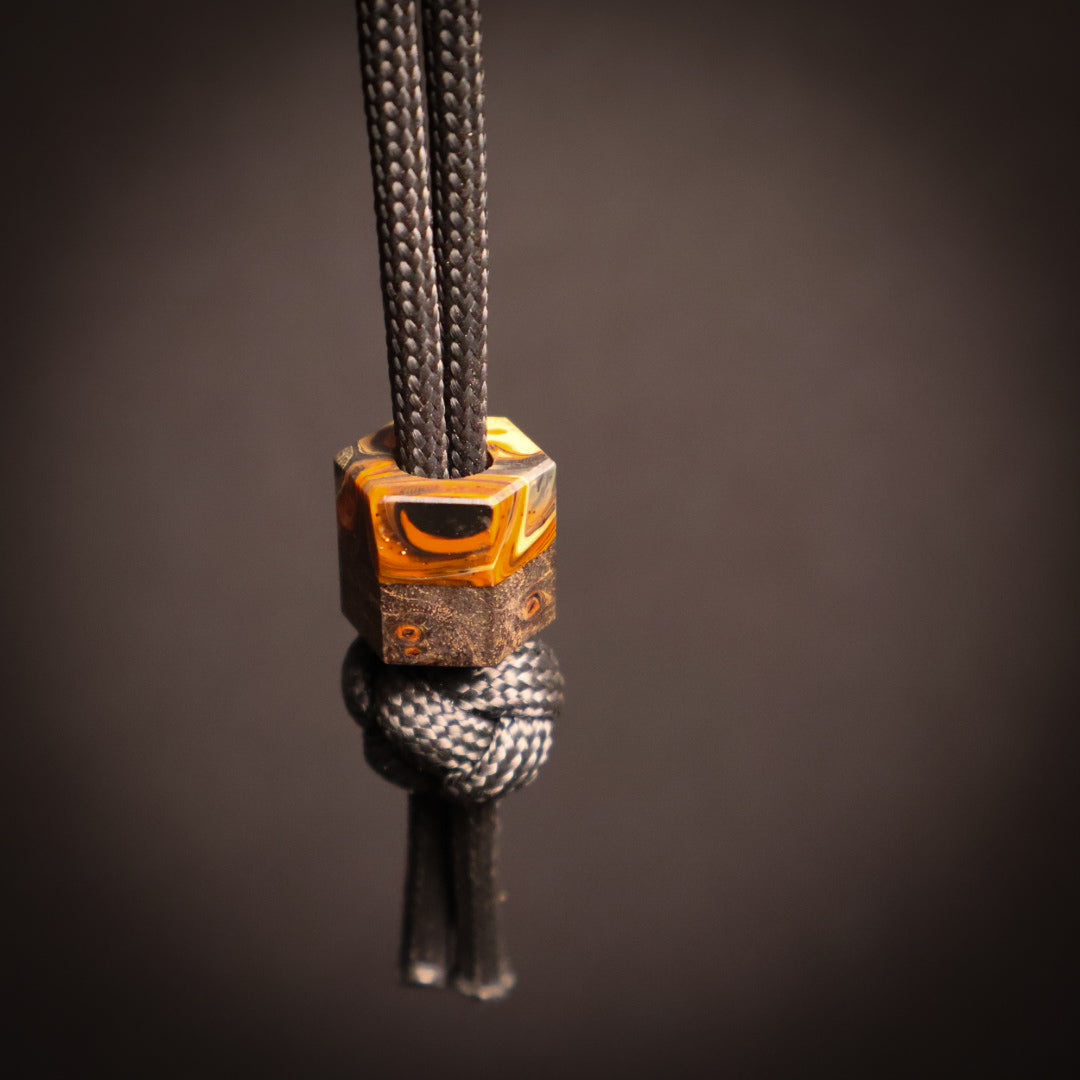 A close-up of a black paracord necklace or lanyard featuring a decorative geometric bead with light and dark brown marbled patterns, tied in a knot below.