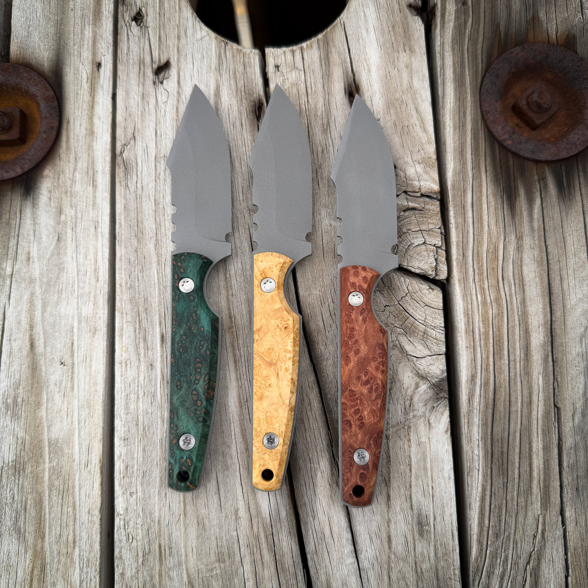 Three knives laying on their sides. All with wood handles of different colors, red, green, and light tan. They are all sitting on a rustic wooden surface 