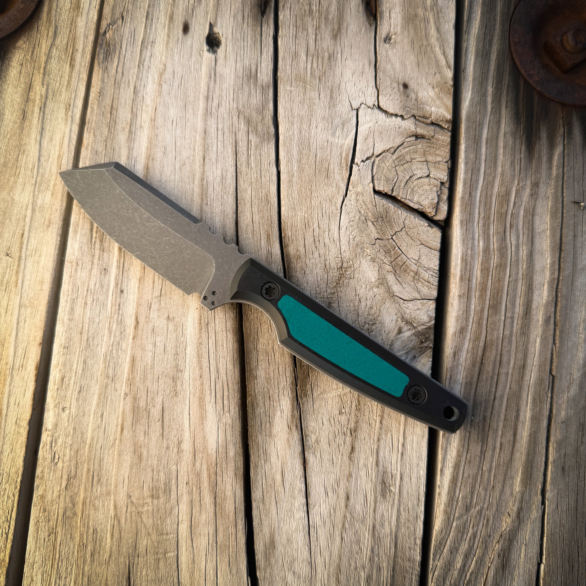 A fixed-blade knife with a matte gray blade and a black handle featuring a prominent teal inlay, resting diagonally on a rustic wooden plank surface.