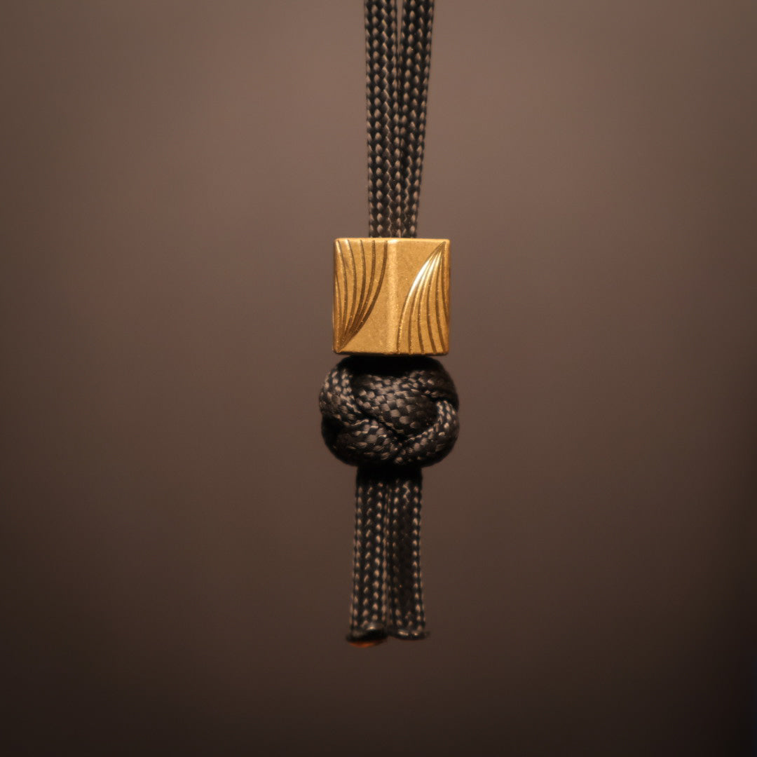 Hexagonal brass lanyard bead with swooped lines milled onto flats. Hanging on black paracord lanyard 