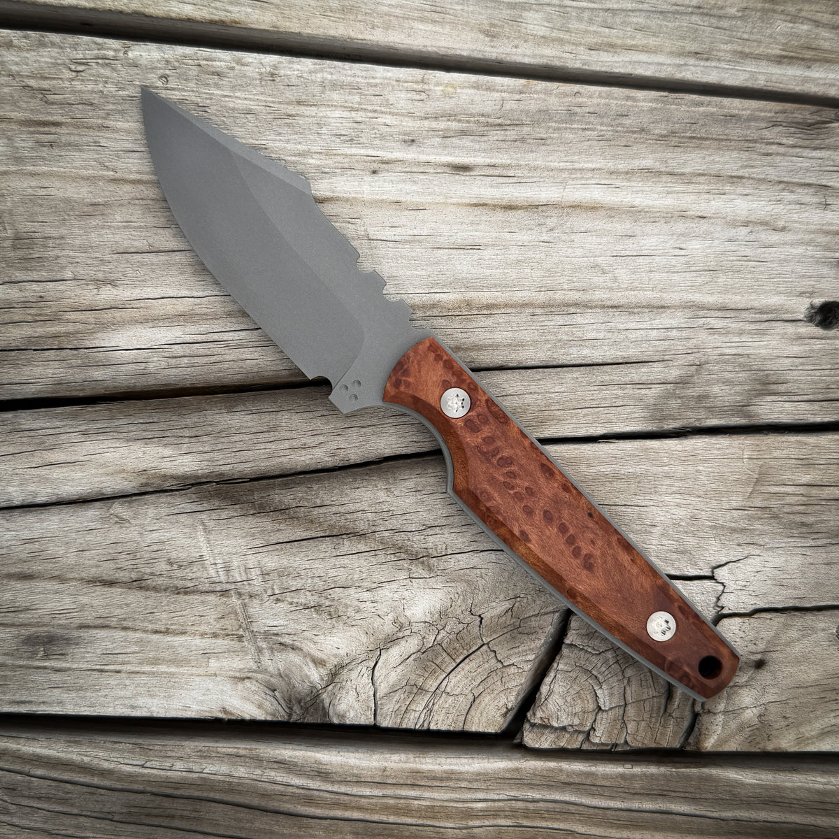 A fixed-blade knife with a matte gray steel blade and a red brown burl wood handle, displayed diagonally on a rustic weathered wood plank surface.