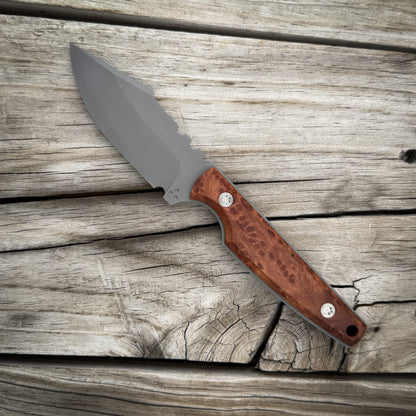 A fixed-blade knife with a matte gray steel blade and a red brown burl wood handle, displayed diagonally on a rustic weathered wood plank surface.