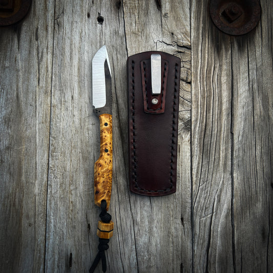 Fixed blade knife with yellow wooden handle sitting on wooden surface next to mahogany leather pocket sheath