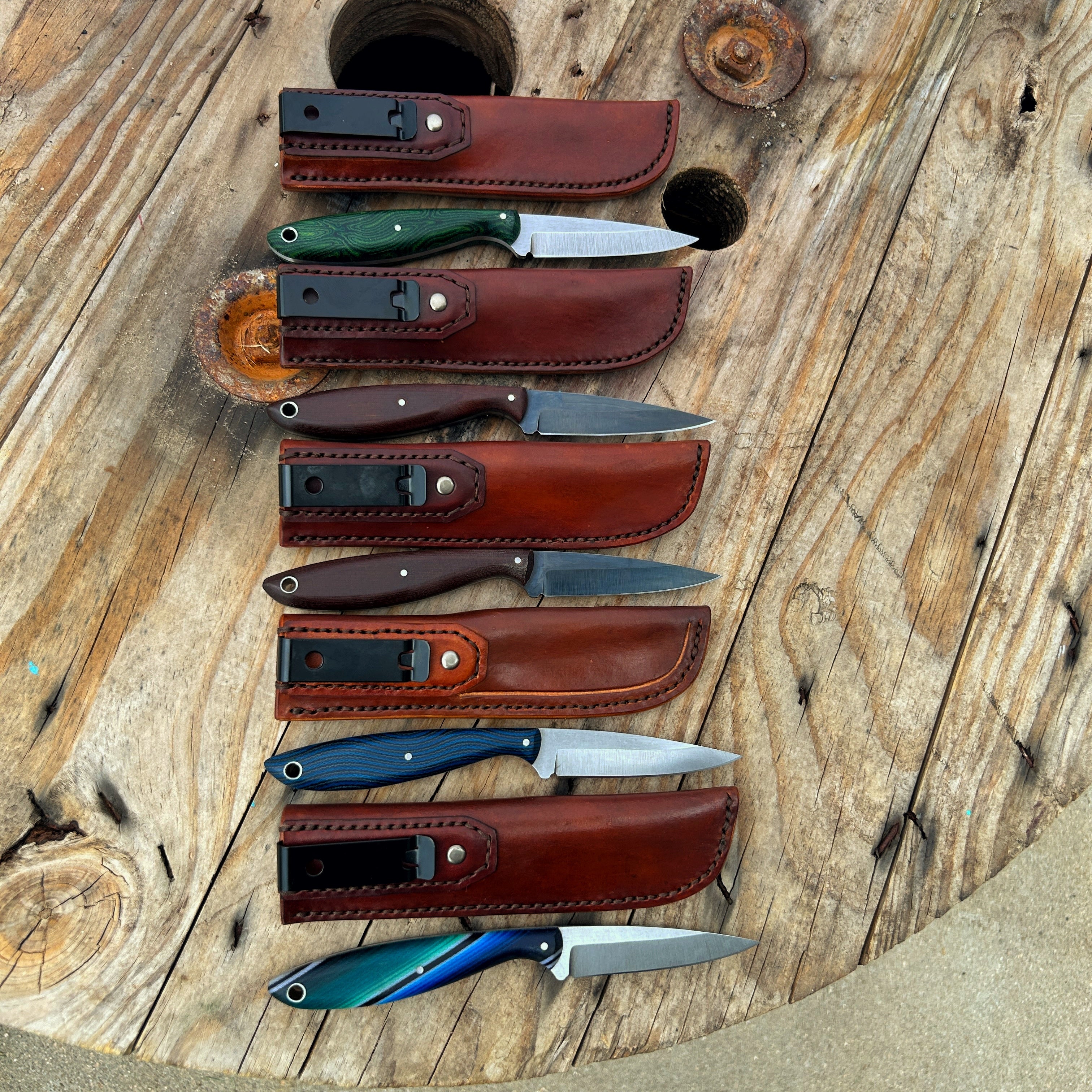 five small knives with leather sheaths sitting on wooden surface 
