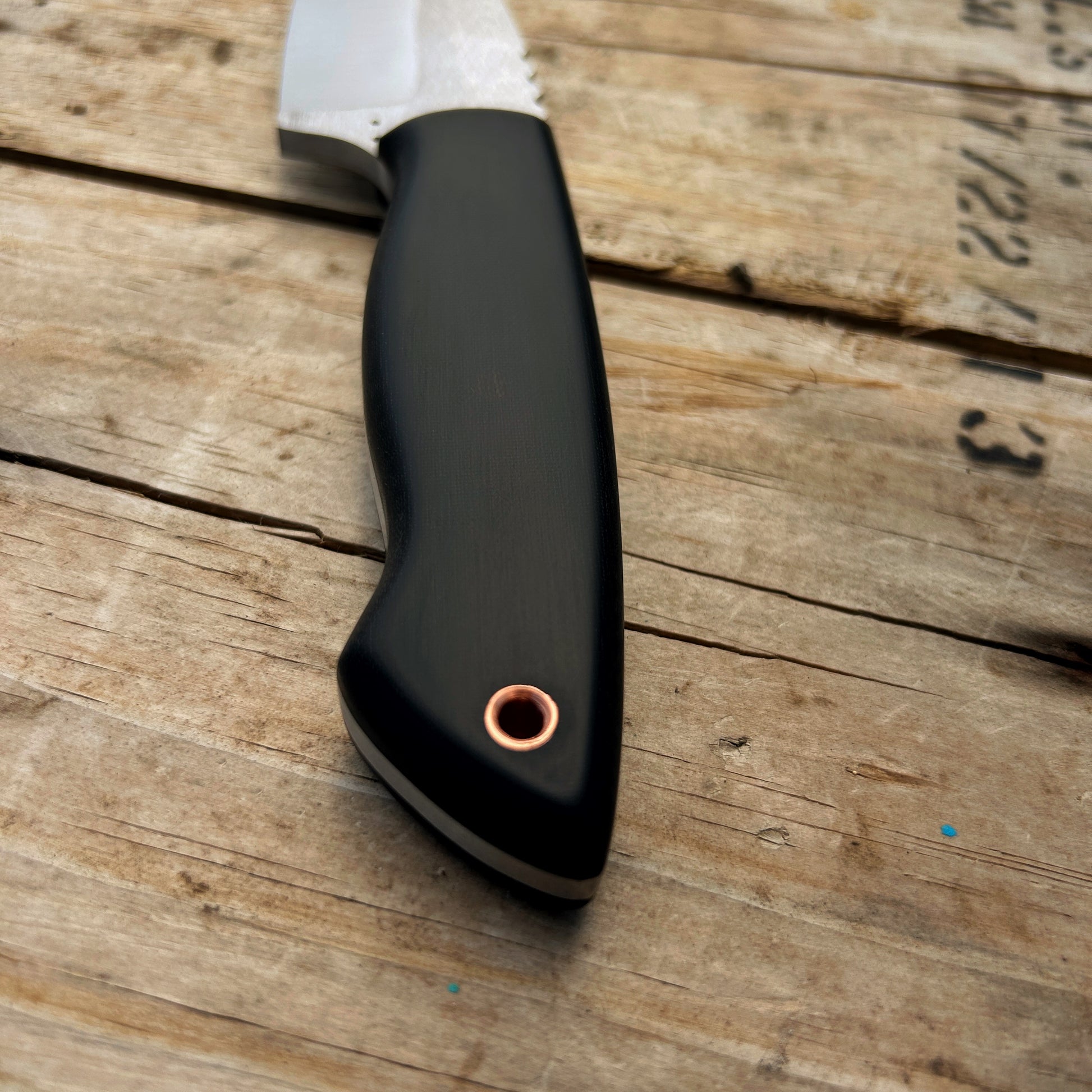 Closeup shot of the handle of the Colorado Hunter knife. The handle is a smooth black linen micarta with black micarta pins & a copper lanyard hole, centered on the handle towards the butt end. The black micarta pins are almost invisible amongst the black handle material. The handle has smooth chamfers leaving a comfortable grip in the hand. 