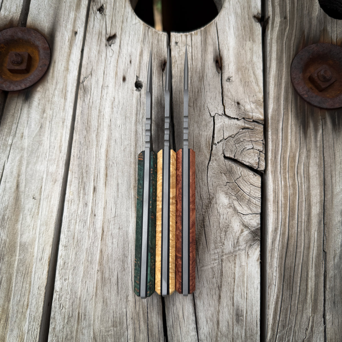 A set of three knives with colorful wood-grain handles, displayed vertically on a rustic weathered wooden plank background.