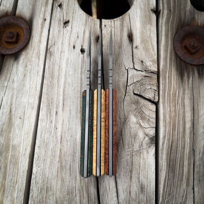 A set of three knives with colorful wood-grain handles, displayed vertically on a rustic weathered wooden plank background.
