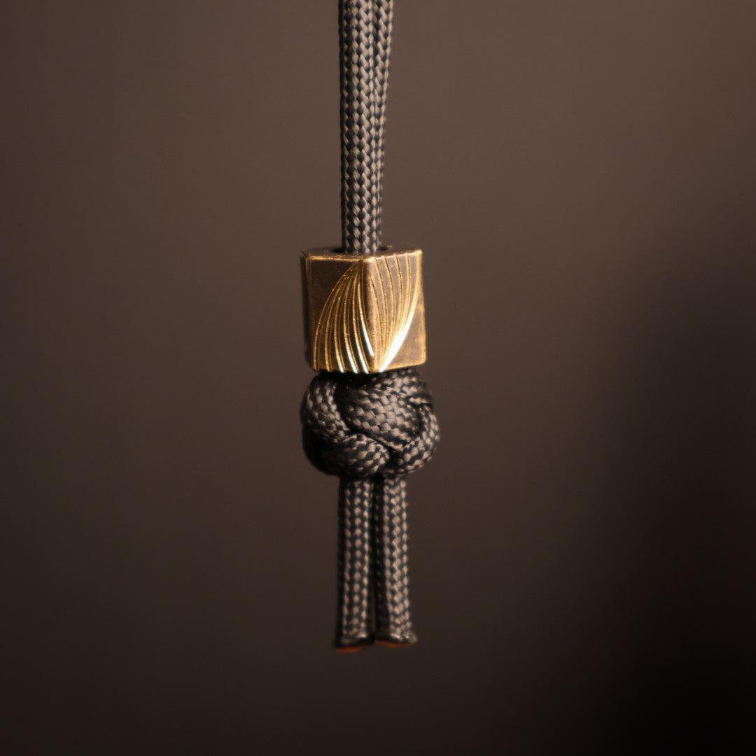 Hexagonal lanyard bead with dark surface and shiny bright milled swoops on the faces hanging from black paracord 