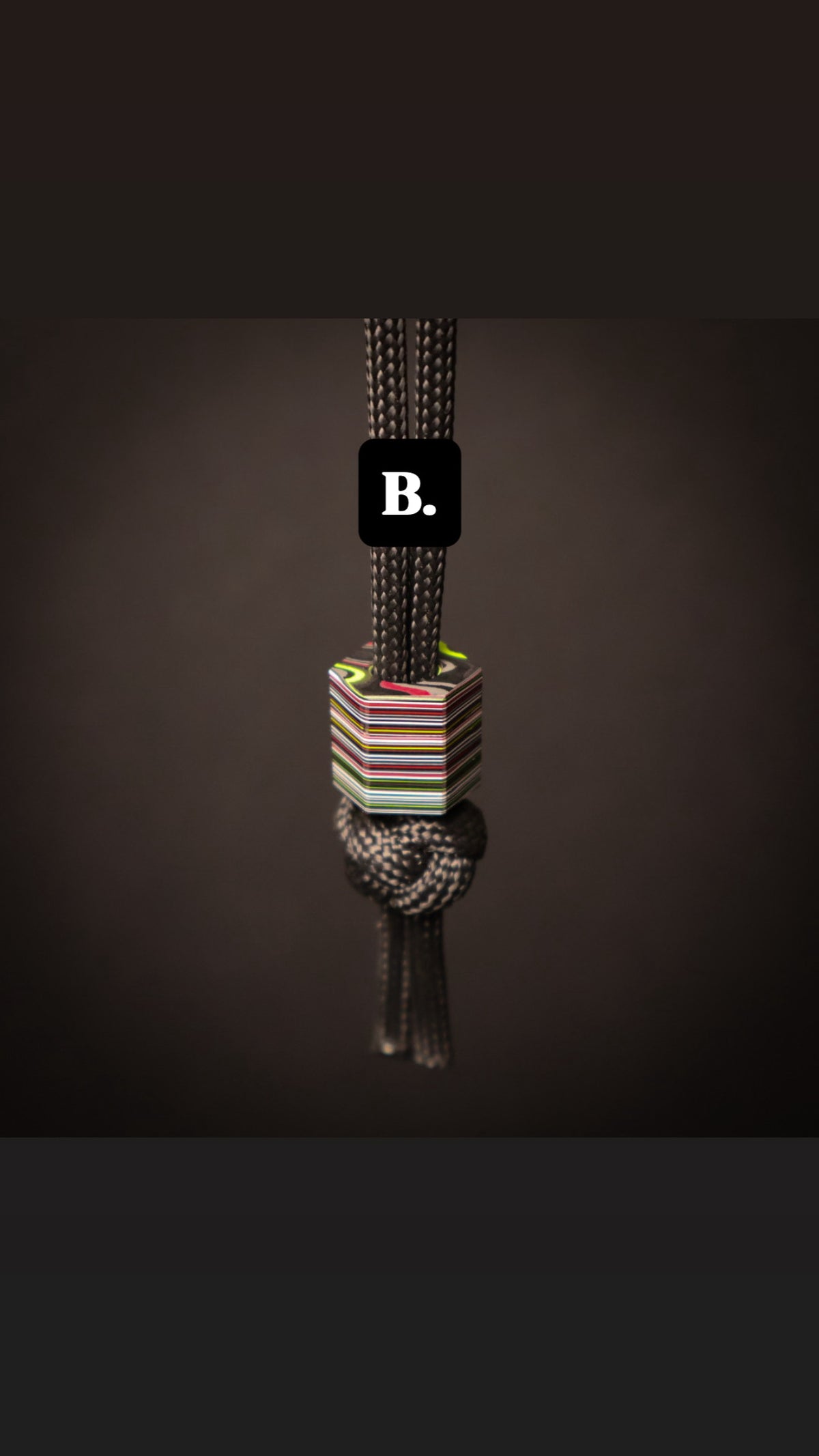A close-up of a braided cord necklace or accessory with a knotted section and a small striped geometric bead, shown against a dark brown background. A black label with the letter 'B.