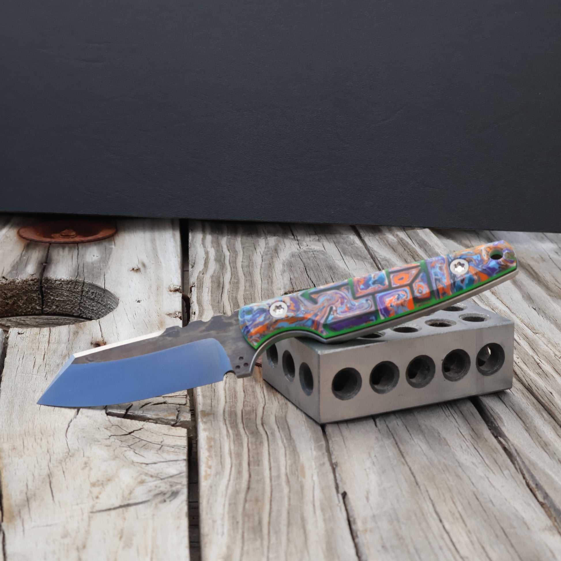 Fixed blade knife with two tone grinds, colorful resin handle resting on metal block with black background