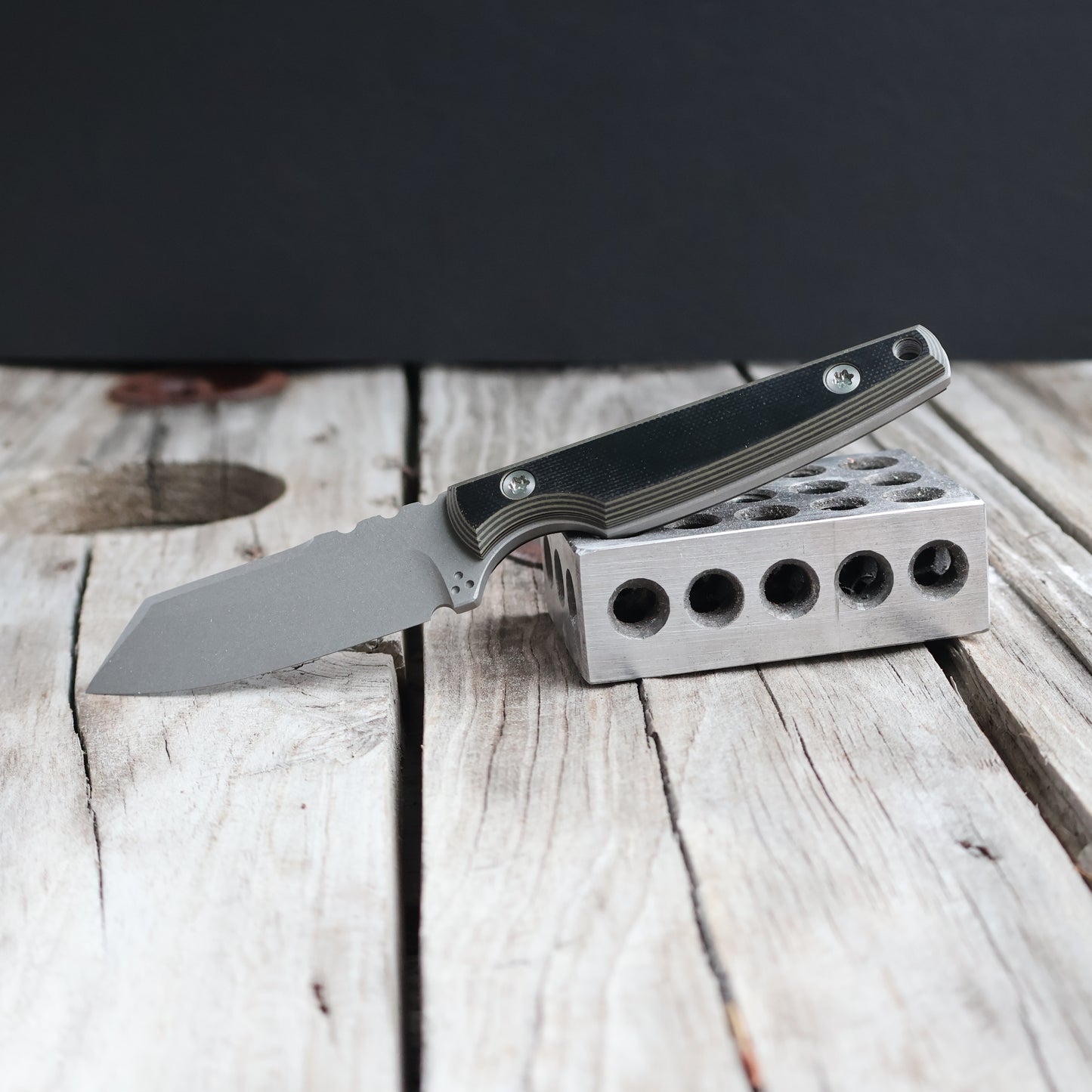 fixed blade knife with dark handle resting on metal block & wooden surface