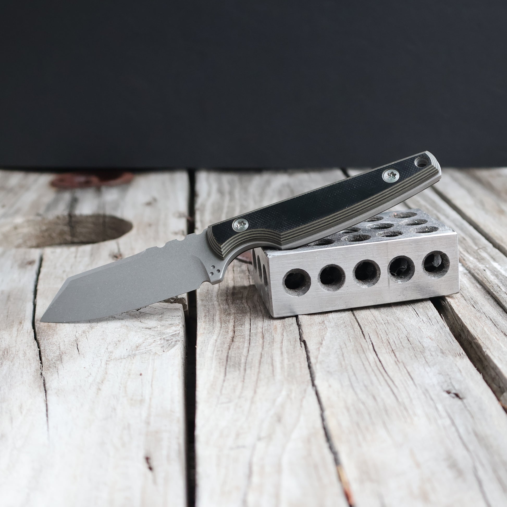 fixed blade knife with dark handle resting on metal block & wooden surface