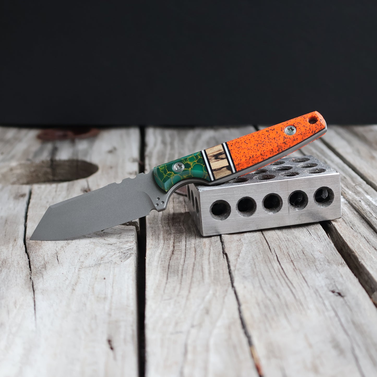 Fixed blade knife with green, tan, and orange combo scale handle sitting on wood surface 