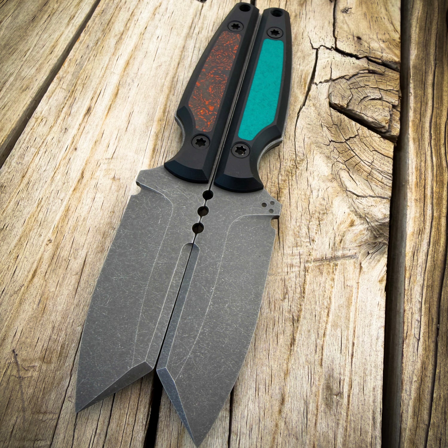 Pair of fixed blade knives sitting spine to spine on wooden surface. Knives have gray blades with black handles with colorful teal and orange gray inlays.