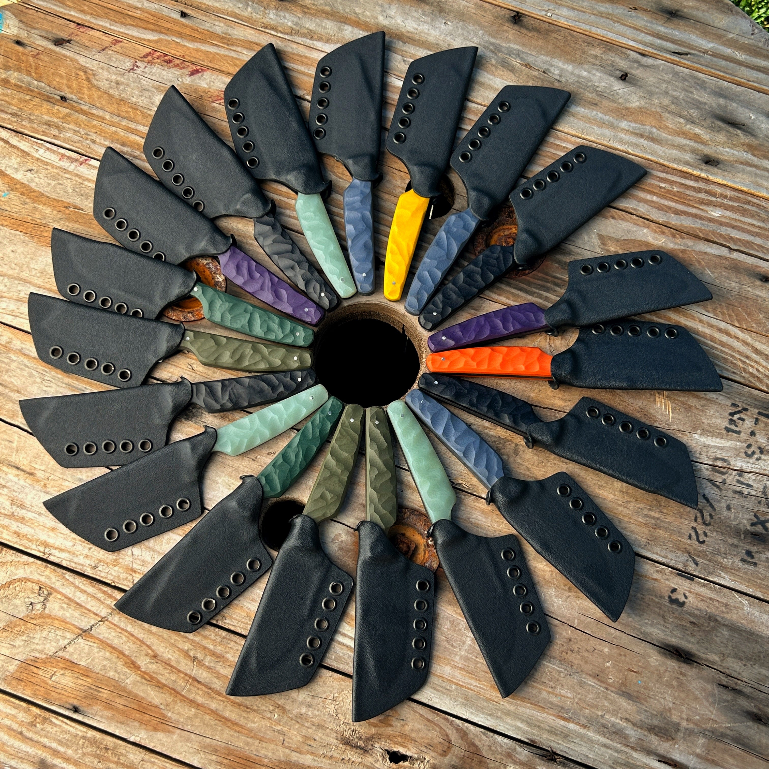 a bunch of knives with kydex sheaths laying on wooden surface in a circle. 