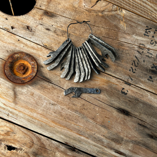 A number of the forge finish keychain bottle openers. All but one threaded on a metal wire demonstrating the attachment method onto a keychain. One is set below so the side profile is visible. They are small opener approx 2"-2.5" in length and 5/8" wide at widest point. The finish is a hammered forge scale finish with dark grey color. 