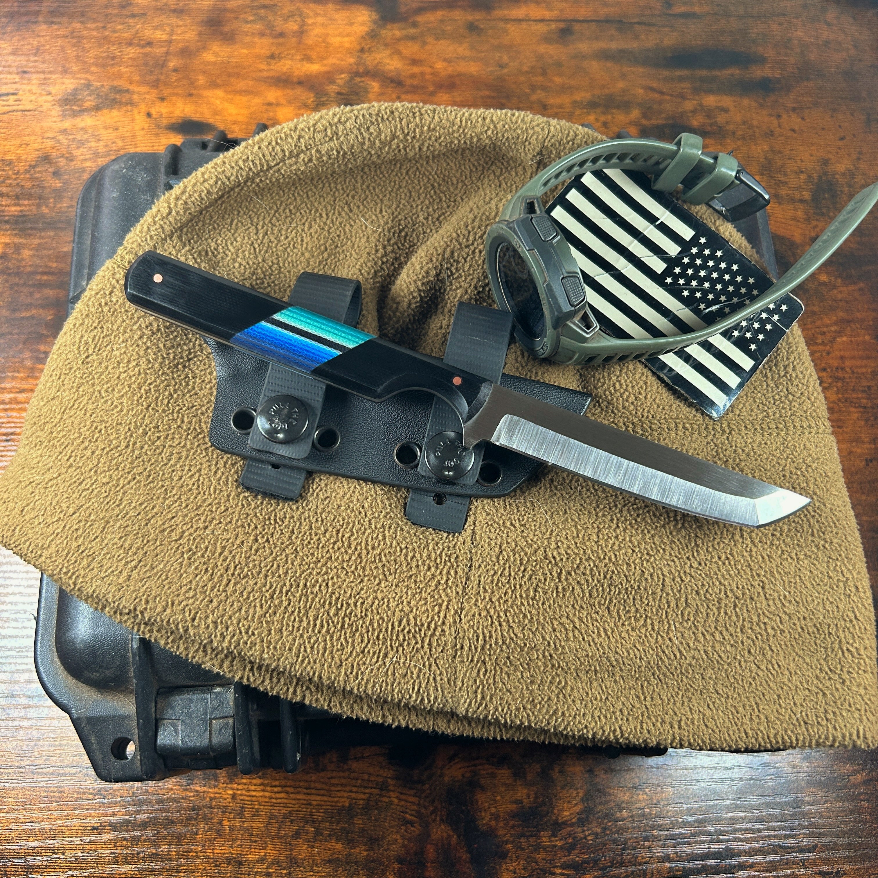A sleek straight tanto blade with a straight handle and one finger groove/cutout. The knife is sitting on top of a kydex sheath with rubberized snap button straps. The knife has a partial flat grind that goes approx 2/3 up the face of the blade. The handle scales are black G-10 with a blue green Mexican blanket micarta segment angled in the middle of the handle. There is one pin at either end of the handle centered and inset from the ends approx 0.5” 