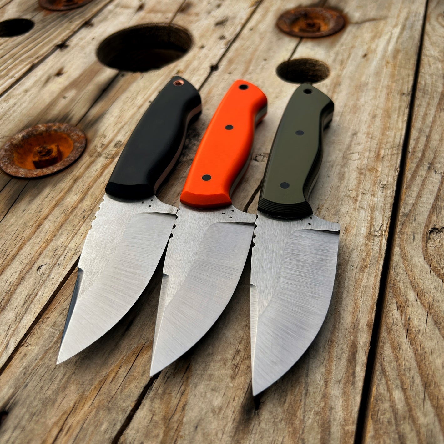 Three of the Colorado Hunter knives with the blade facing the camera and handles away. There is a black, orange, & OD green/black handled version pictured. The blades exhibit three dots in a triangle (makers mark) just behind the plunge line. 