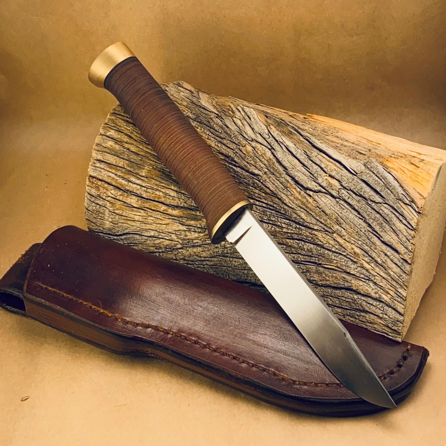 Classic styled (think your grandpas hunting knife)skinny hunting/fishing knife with a false edge at the tip. It has a thing brass plate guard, stacked brown leather handle, & a half inch thick swelled brass pommel. It is leaned up against a weathered piece of firewood with a dark brown leather belt sheath laying underneath it.